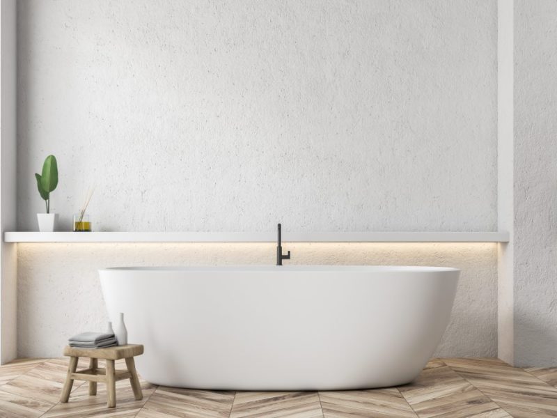 Interior of modern bathroom with white walls, wooden floor and white bathtub standing next to a shelf on the wall.